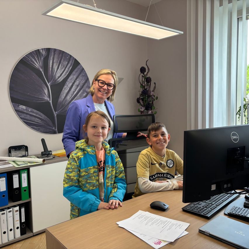 Eine Frau steht hinter einem Schreibtisch mit zwei Kindern vor einem Computer. Die Kinder lächeln. An der Wand befindet sich ein großes schwarzes kreisförmiges Design.