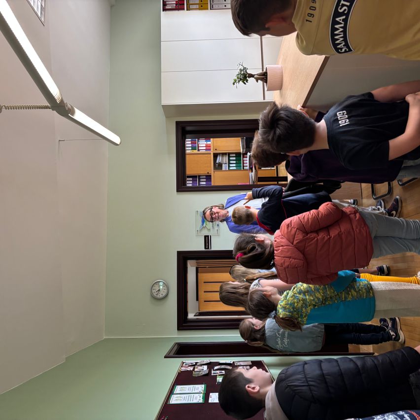A group of children and a teacher gather in a classroom, observing a teacher who is explaining something. There are cabinets, a clock, and posters on the walls.