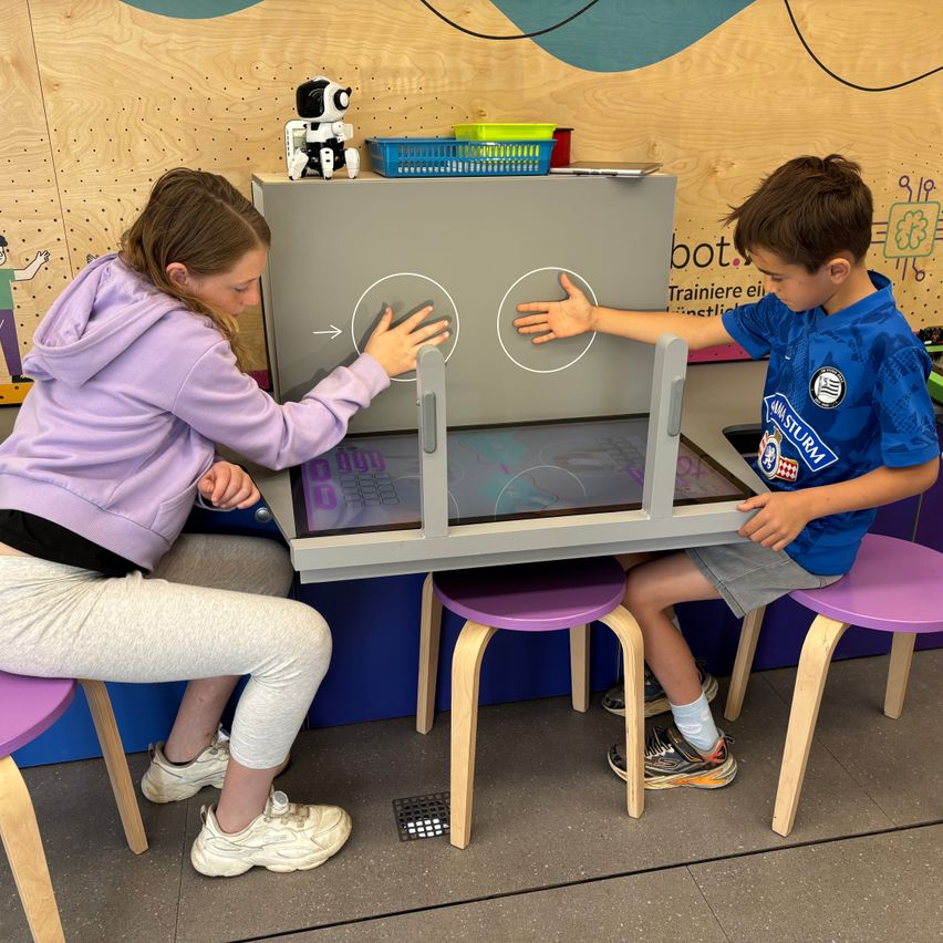 Ein Mädchen und ein Junge sitzen auf Hockern und interagieren mit einem Touchscreen. Das Mädchen trägt eine lila Hoodie, und der Junge trägt ein blaues Shirt. Hinter ihnen ist eine Holzwand mit einem Roboter.