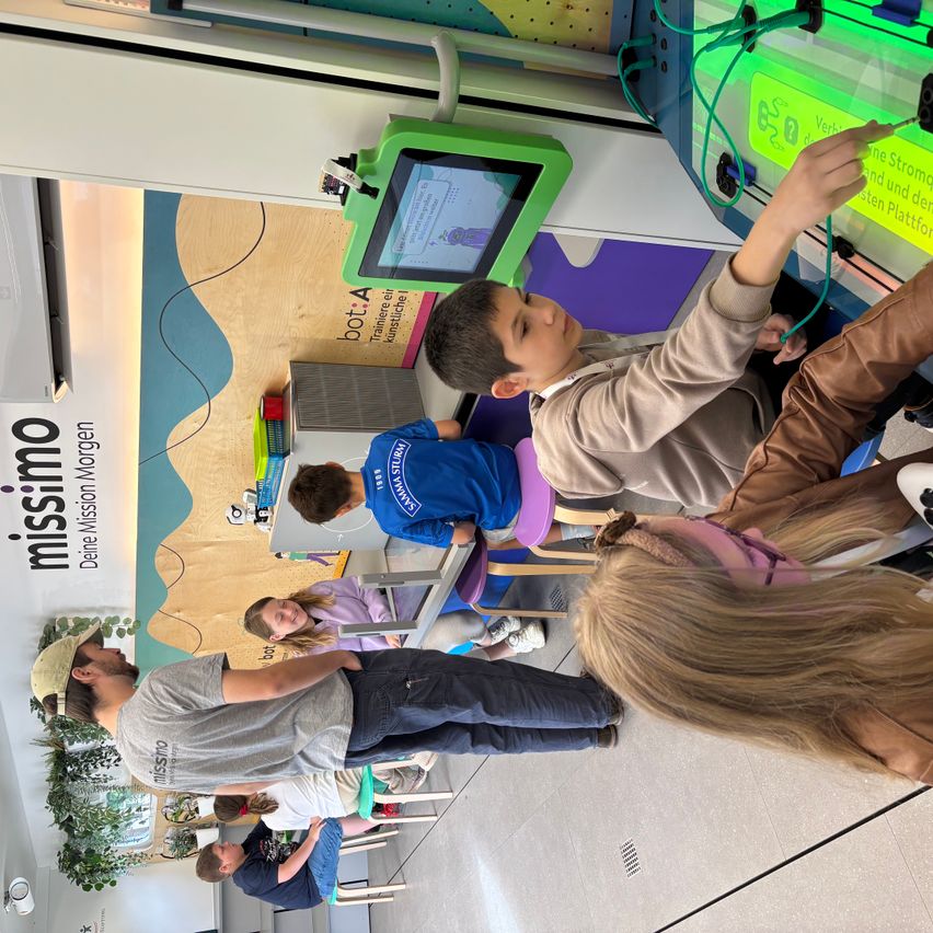Eine Gruppe von Kindern versammelt sich um eine interaktive Maschine. Sie sind mit dem Gerät beschäftigt, ein Kind zeigt auf den Bildschirm. Der Raum hat ein modernes Design mit einem farbenfrohen Hintergrund.