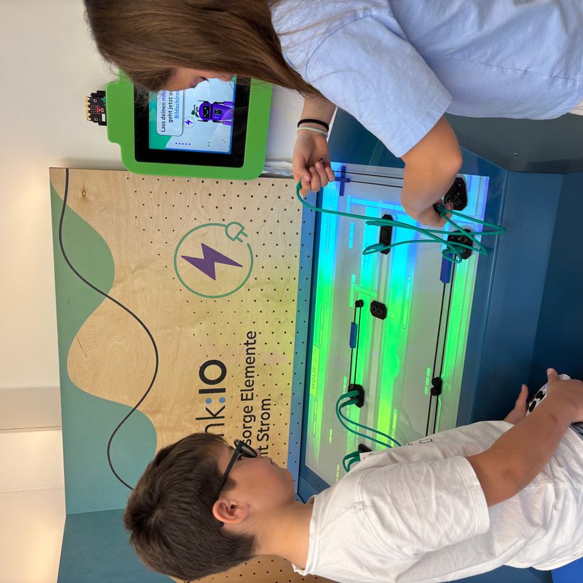 Zwei Kinder spielen mit einer grünen, interaktiven elektrischen Tafel. Ein Kind hält die Tafel, während das andere sie aufmerksam betrachtet. Die Tafel hat einen Bildschirm und verschiedene farbige Lichter.