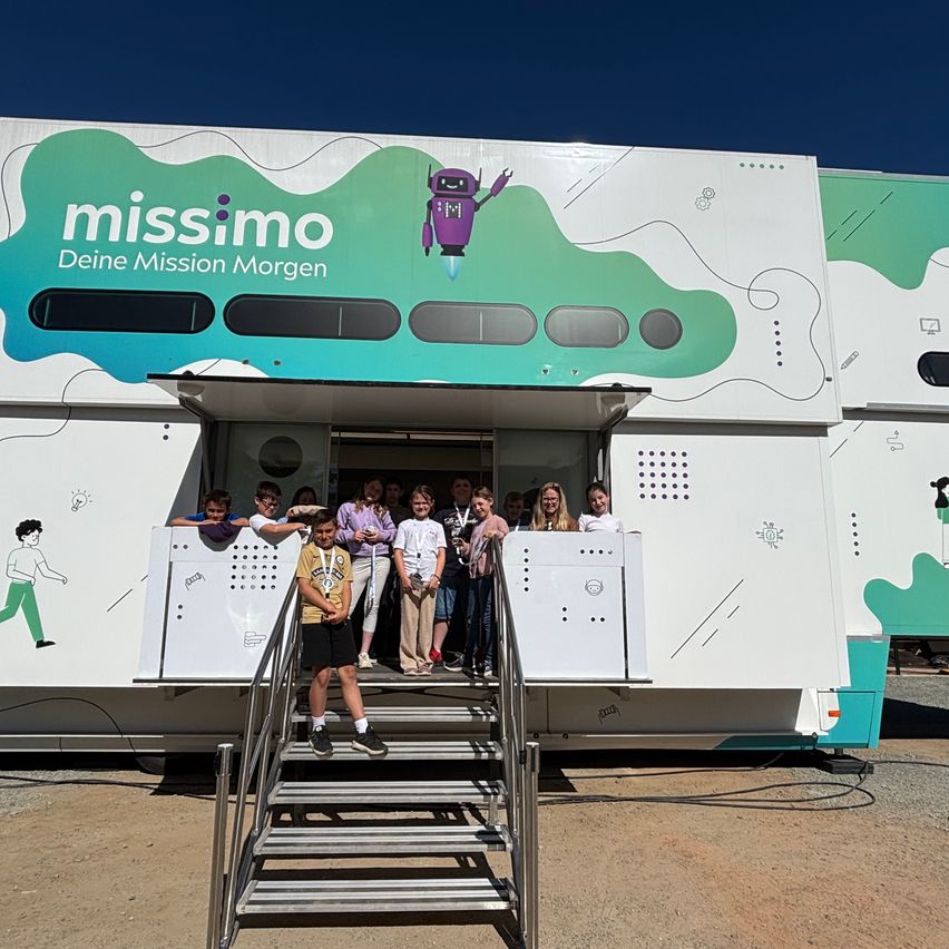 Kinder erkunden ein mobiles Bildungsfahrzeug namens Missimo. Das Fahrzeug ist mit farbenfrohen Grafiken bemalt und hat ein Raketenlogo. Es ist im Freien geparkt.
