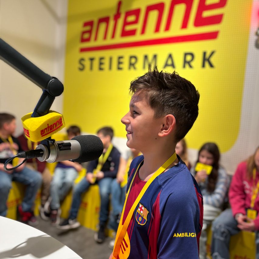 Ein junger Junge in einem FC Barcelona-Trikot spricht in ein Mikrofon vor einem gelben Antennen-Steiermark-Schild, während andere Kinder auf gelben Bänken hinter ihm sitzen.