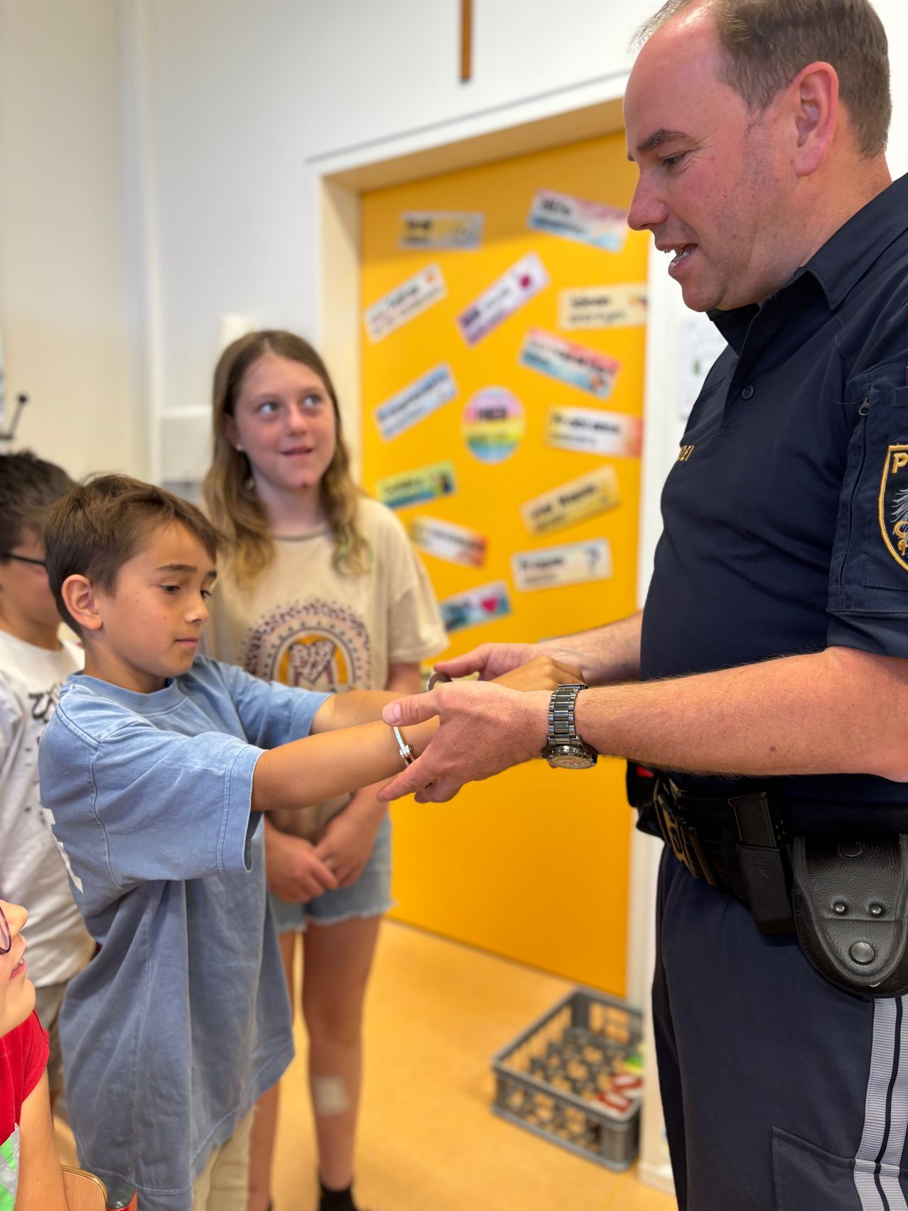 Ein Polizeibeamter zeigt einem jungen Jungen ein Armband, während ein Mädchen hinter ihnen zusieht. Der Klassenraum ist mit verschiedenen Aufklebern an einer gelben Wand dekoriert.