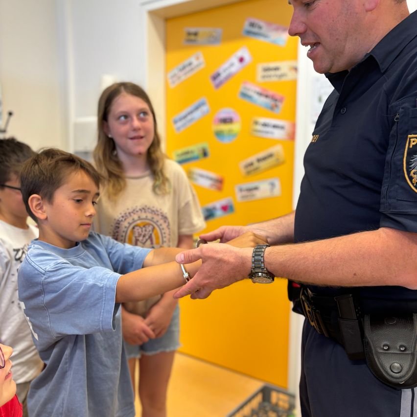 Ein Polizeibeamter zeigt einem jungen Jungen ein Armband, während ein Mädchen hinter ihnen zusieht. Der Klassenraum ist mit verschiedenen Aufklebern an einer gelben Wand dekoriert.