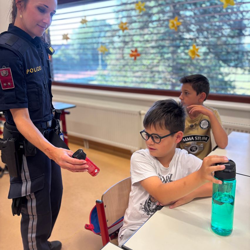 Eine Polizistin zeigt einem Jungen, wie man einen Feuerlöscher benutzt, während ein anderer Junge zusieht. Sie befinden sich in einem Klassenzimmer.