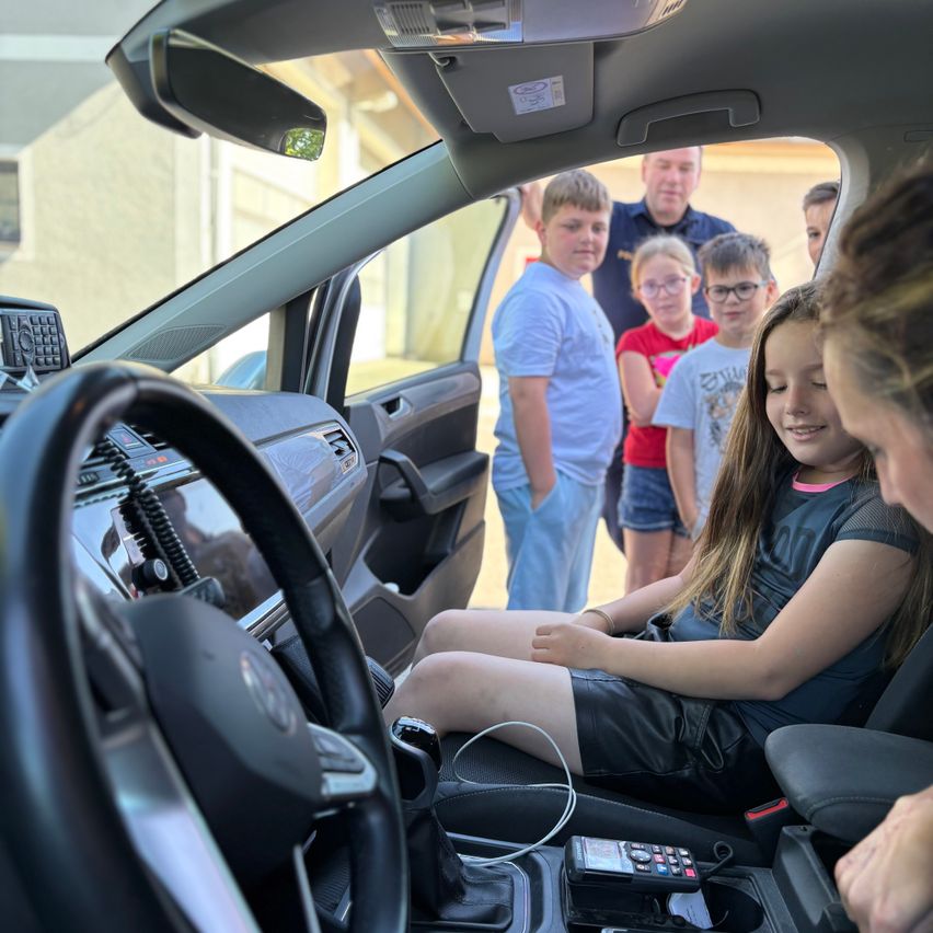Ein Mädchen sitzt auf dem Fahrersitz eines Autos, während eine Frau ihre Arbeit überprüft. Hinter ihr stehen eine Gruppe von Kindern und Erwachsenen bei der Autotür und beobachten.