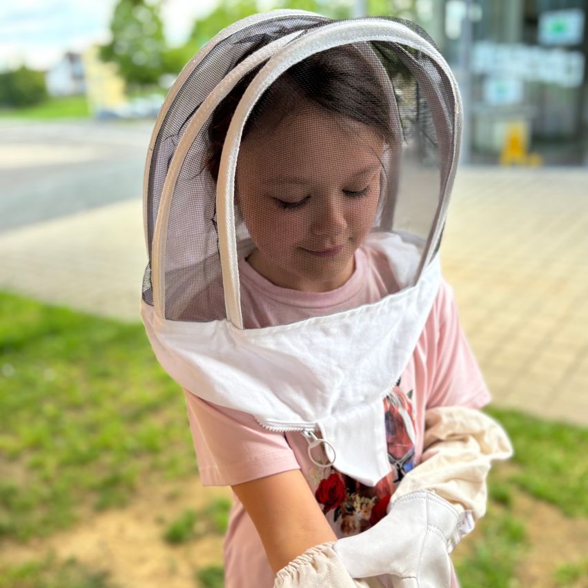 Ein kleines Mädchen mit einem rosa T-Shirt und weißer Bienenkleidung schaut nach unten. Sie hat ein Bienenschutznetz auf dem Kopf.