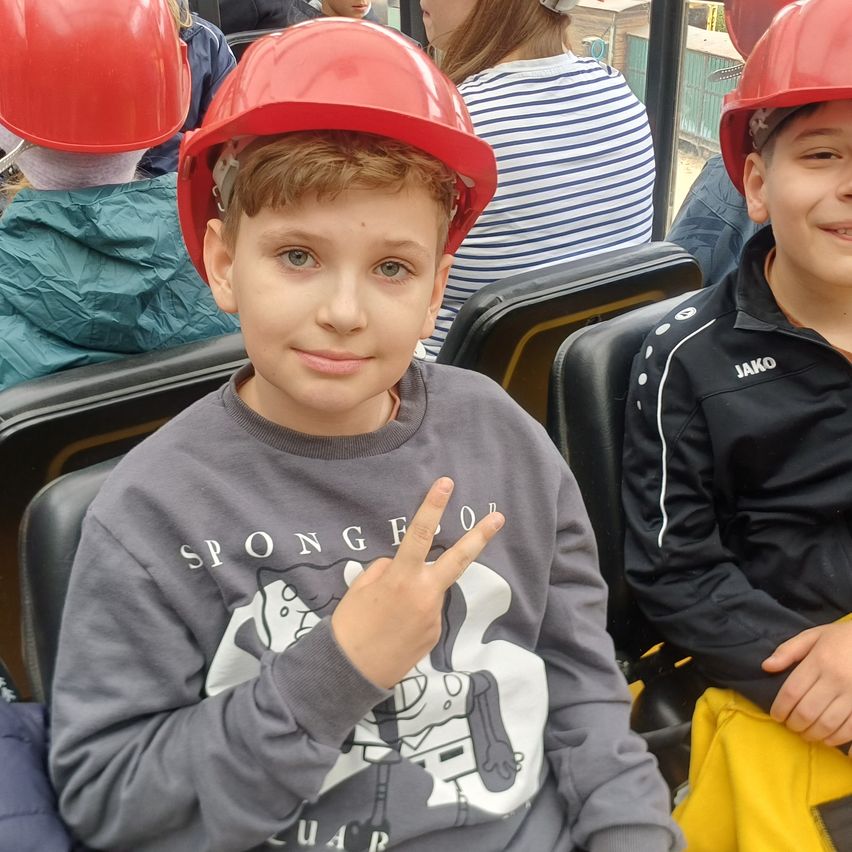 Ein Junge in einem grauen Shirt und rotem Helm sitzt auf einem Stuhl mit lächelndem Gesicht. Andere Kinder in roten Helmen sitzen hinter ihm.