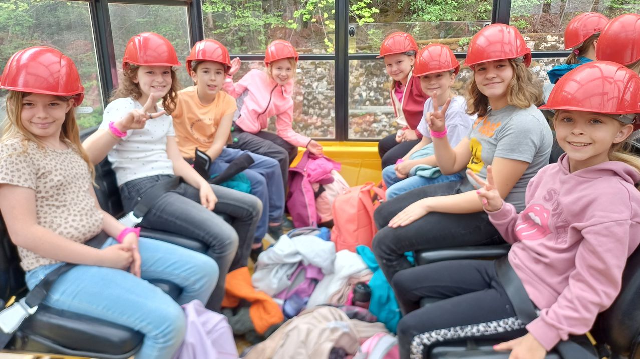 Eine Gruppe von Mädchen mit roten Helmen sitzt in einem Bus, lächelt und hat verschiedene Rucksäcke und Gegenstände um sich herum verstreut.