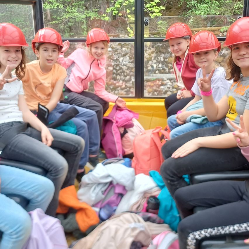Eine Gruppe von Mädchen mit roten Helmen sitzt in einem Bus, lächelt und hat verschiedene Rucksäcke und Gegenstände um sich herum verstreut.