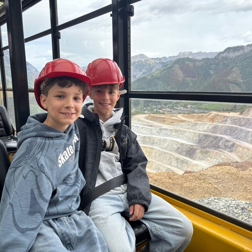 Zwei junge Jungen mit roten Schutzhelmen sitzen in einem Fahrzeug, wahrscheinlich einer Minentour, und blicken aus einem Fenster auf eine bergige Bergbaulandschaft.