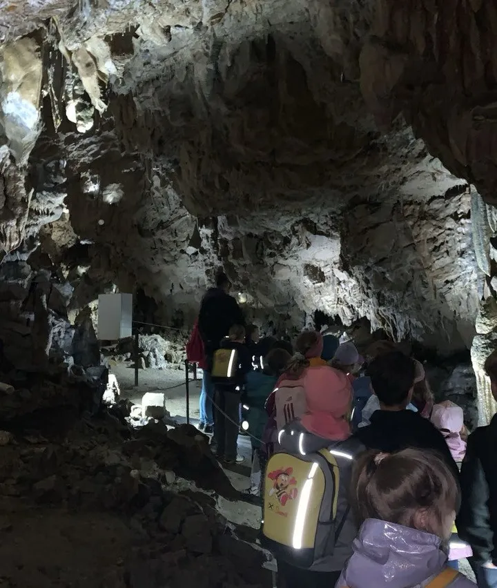 Eine Gruppe von Menschen, einschließlich Kindern, erkundet eine Höhle. Sie sind mit Rucksäcken und Mützen ausgestattet. Die Höhle hat felsige Wände und ist schwach beleuchtet.