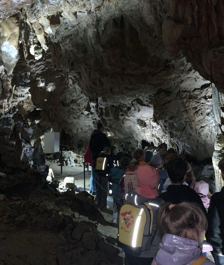 Eine Gruppe von Menschen, einschließlich Kindern, erkundet eine Höhle. Sie sind mit Rucksäcken und Mützen ausgestattet. Die Höhle hat felsige Wände und ist schwach beleuchtet.