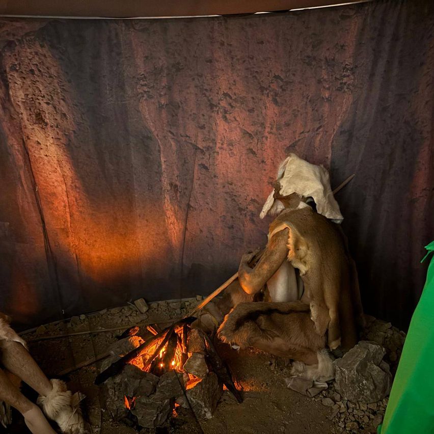 Eine Person sitzt in einer Höhle neben einem Feuer mit beleuchtetem Hintergrund. Die Figur ist in Tierhäute gekleidet und hält einen Speer.