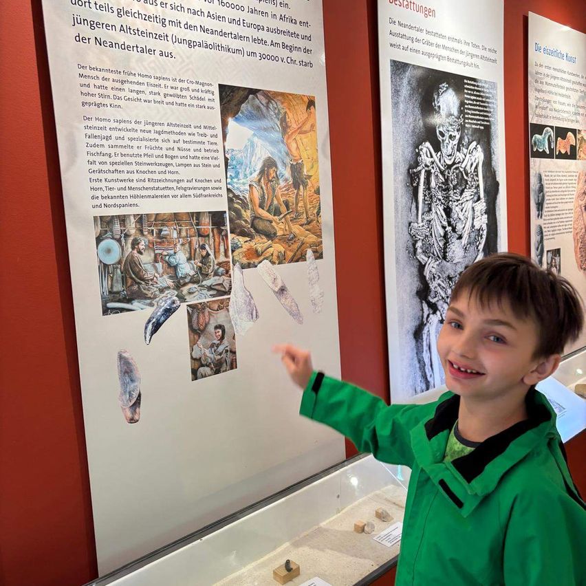 Ein junger Junge lächelt und zeigt auf eine Ausstellung in einem Museum. Die Ausstellung hat Text und Bilder von prähistorischem Leben. Der Junge trägt eine grüne Jacke und ist von Glasvitrinen mit Artefakten umgeben.