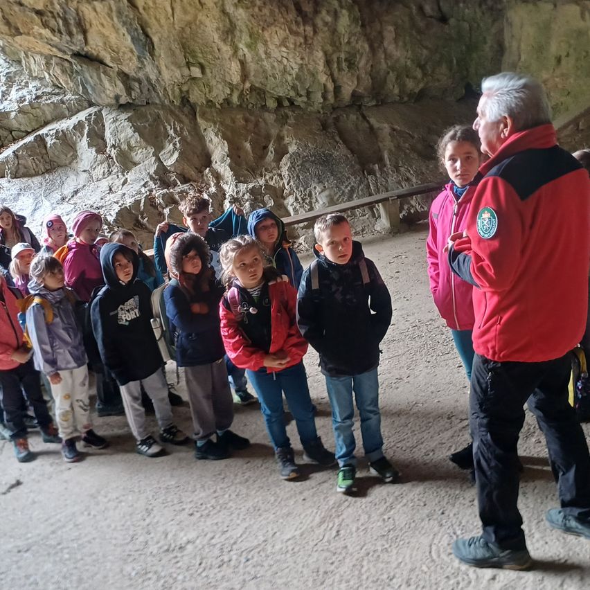 Eine Gruppe von Kindern steht in einer Höhle mit einem Mann in einem roten Jacken, der eine Führung gibt. Die Kinder tragen Jacken und Turnschuhe und stehen in einem Halbkreis.