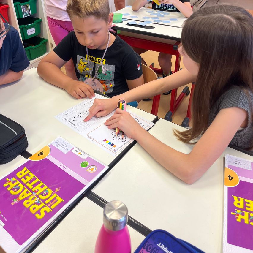 Kinder arbeiten an einer Klassenaktivität, ein Junge füllt eine Arbeitsmappe aus, während ein Mädchen ihm hilft. Sie sind von verschiedenen Schulmaterialien umgeben.
