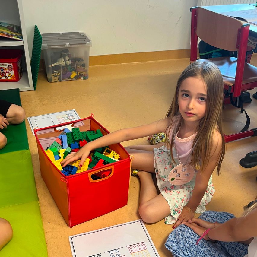 Ein junges Mädchen sitzt auf dem Boden und spielt mit bunten Bausteinen aus einer roten Aufbewahrungsbox. Sie trägt ein rosa Oberteil und eine Namensplakette.
