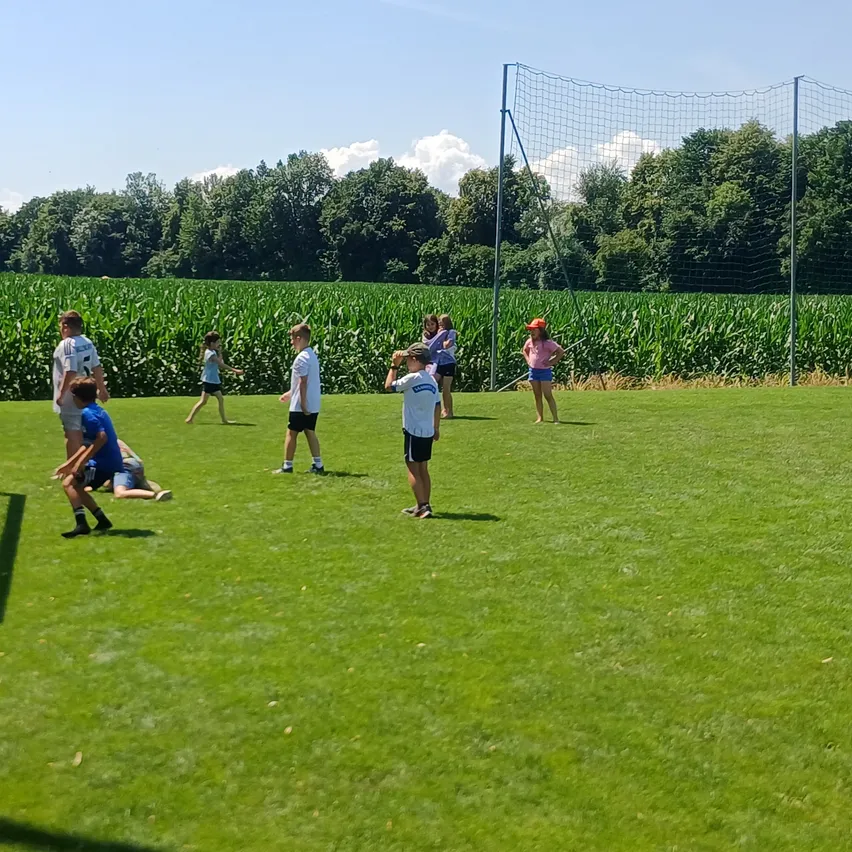 Eine Gruppe von Kindern spielt auf einem Grasfeld mit einem Maisfeld im Hintergrund. Einige tragen Turnschuhe, und ein Torpfosten ist sichtbar. Bäume umgeben das Gebiet.