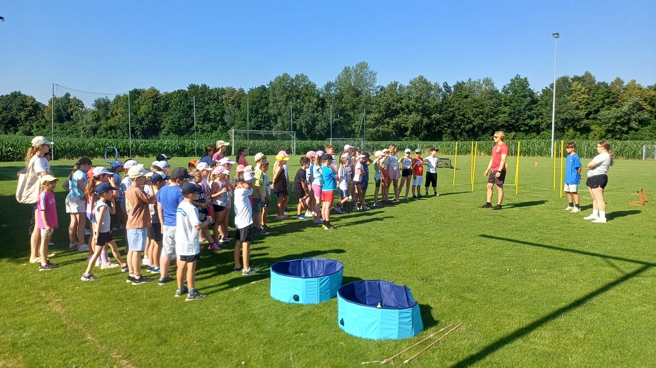 Eine Gruppe von Kindern und Erwachsenen steht auf einem Rasenfeld, trägt Hüte und Turnschuhe. Eine Frau steht vor ihnen und unterrichtet sie wahrscheinlich. Zwei blaue Wannen stehen auf dem Boden. In der Ferne gibt es Bäume und ein Tor.