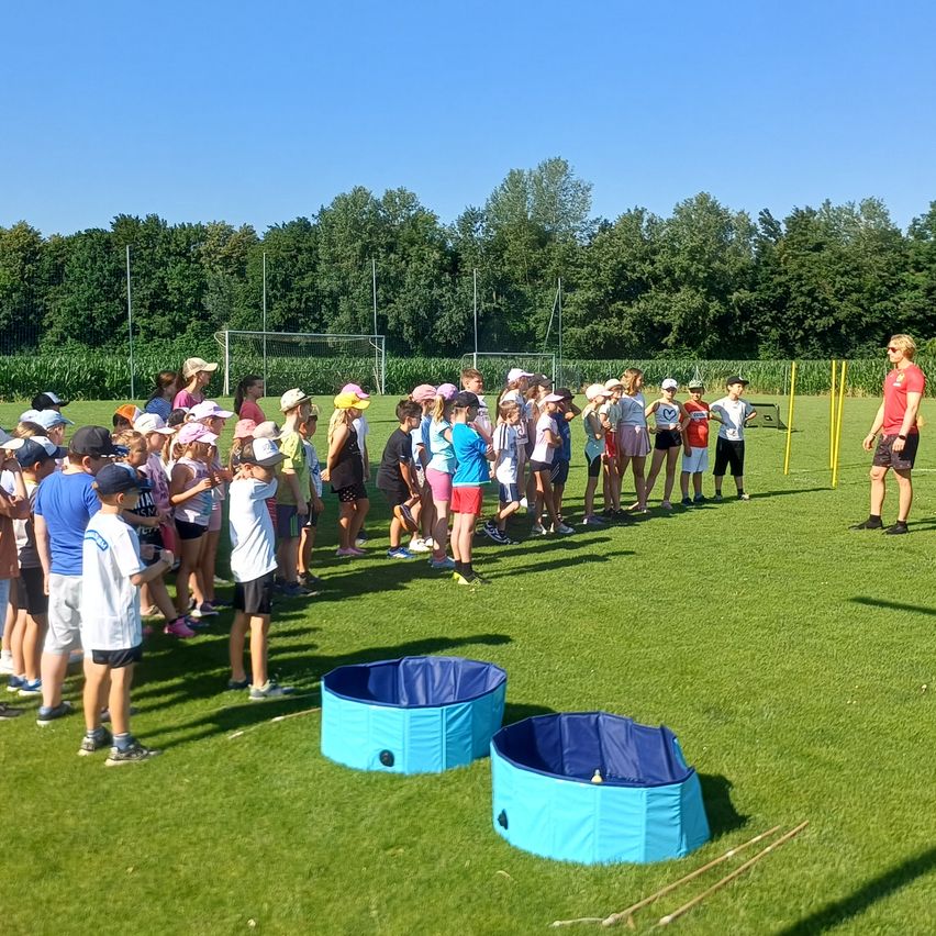 Eine Gruppe von Kindern und Erwachsenen steht auf einem Rasenfeld, trägt Hüte und Turnschuhe. Eine Frau steht vor ihnen und unterrichtet sie wahrscheinlich. Zwei blaue Wannen stehen auf dem Boden. In der Ferne gibt es Bäume und ein Tor.