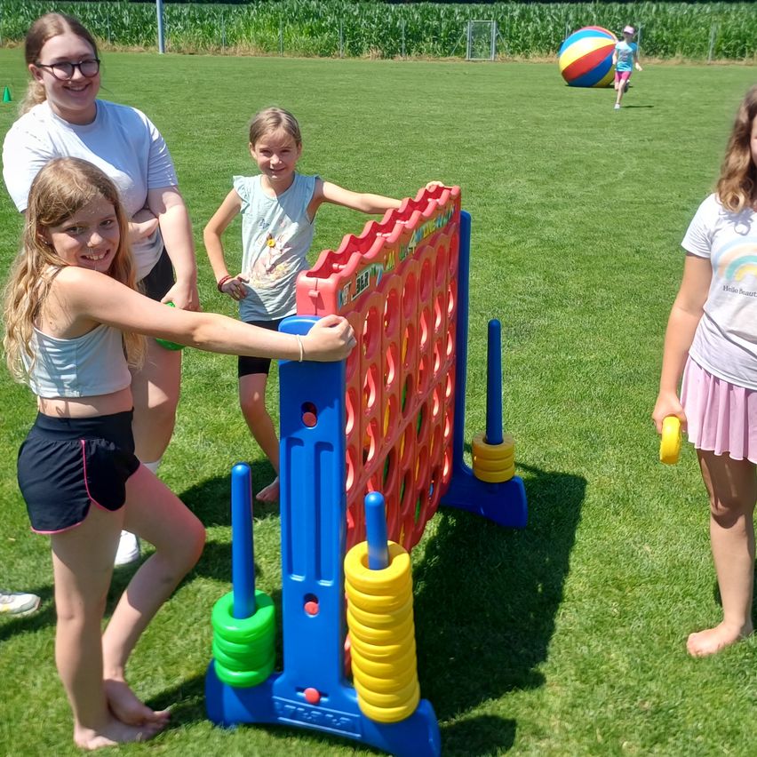 Vier Mädchen spielen draußen an einem sonnigen Tag ein Spiel von Connect Four. Das Spielbrett ist farbenfroh mit einer Mischung aus blauen, roten und gelben Teilen. Ein anderes Kind läuft mit einem bunten Ball.