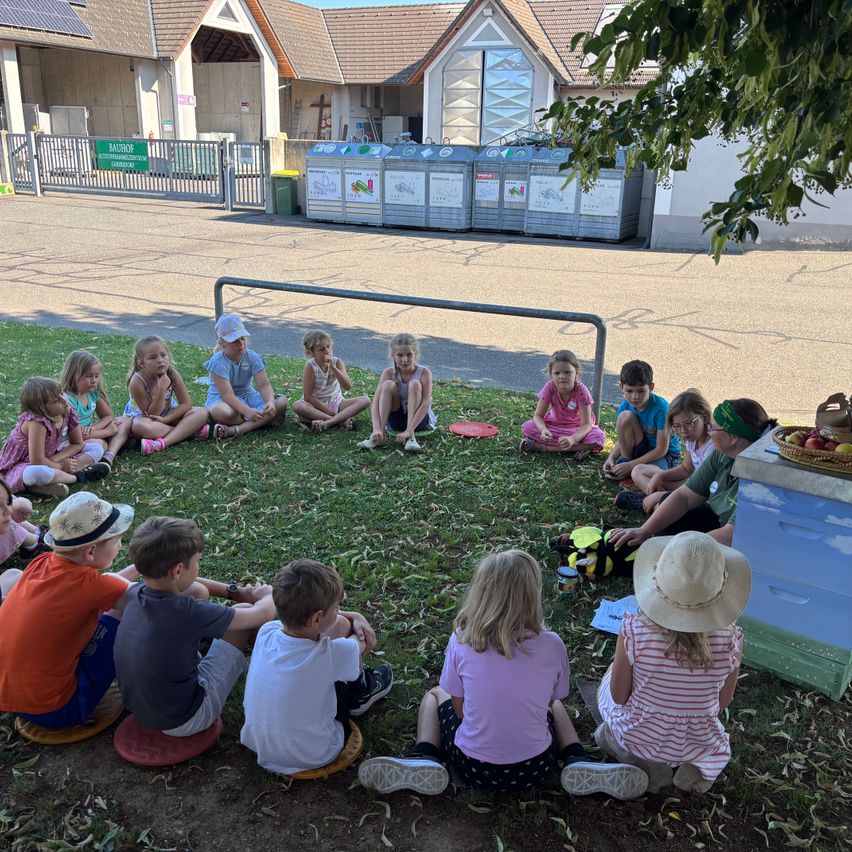 Eine Gruppe von Kindern sitzt im Kreis auf dem Rasen vor einem Gebäude mit Solarmodulen. Ein Korb mit Äpfeln und ein Bienenstock sind in der Nähe.