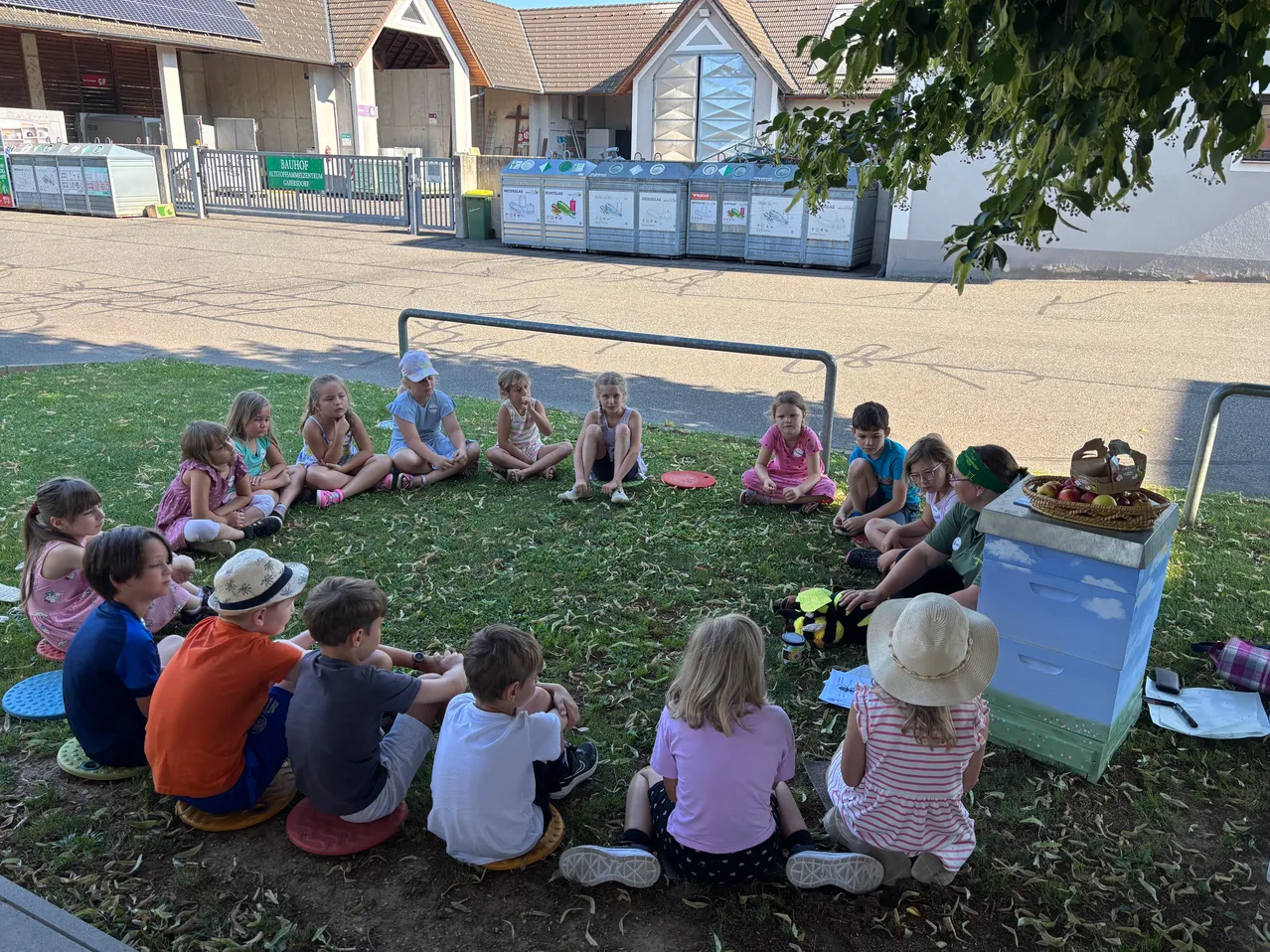 Eine Gruppe von Kindern sitzt im Kreis auf dem Rasen vor einem Gebäude mit Solarmodulen. Ein Korb mit Äpfeln und ein Bienenstock sind in der Nähe.