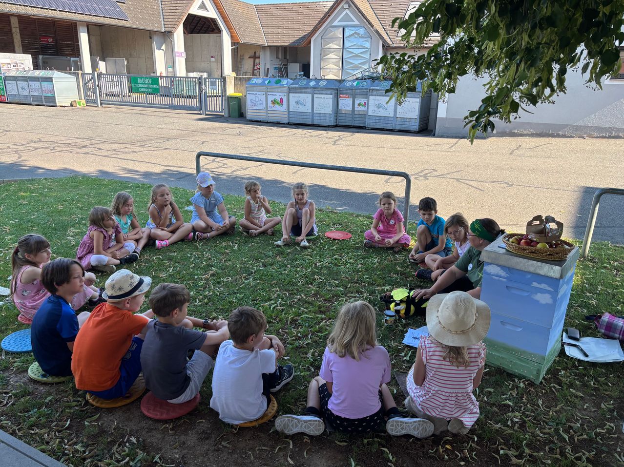 Eine Gruppe von Kindern sitzt im Kreis auf dem Rasen vor einem Gebäude mit Solarmodulen. Ein Korb mit Äpfeln und ein Bienenstock sind in der Nähe.
