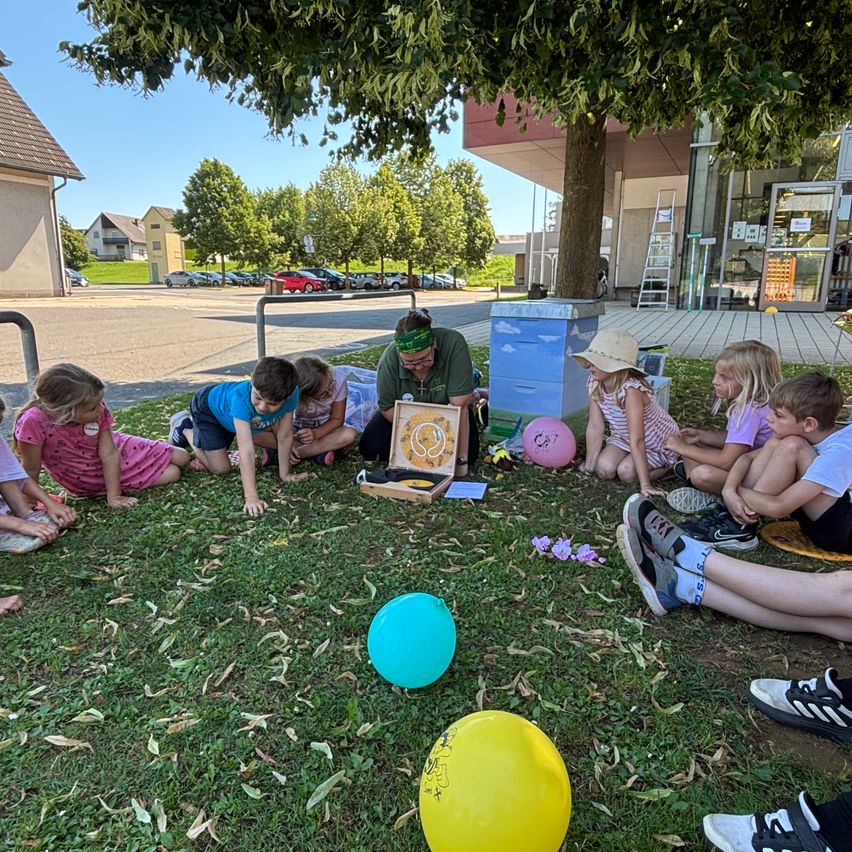 Eine Gruppe von Kindern und einem Mann sitzen im Kreis auf Gras und betrachten eine Schachtel mit einer Uhr. Der Mann trägt ein Stirnband. Es gibt Ballons und Blumen auf dem Boden.