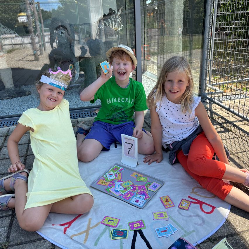 Drei Kinder sitzen auf dem Boden vor einem Brettspiel. Ein Junge hält eine Karte mit der Zahl 7. Sie tragen alle Kronen.