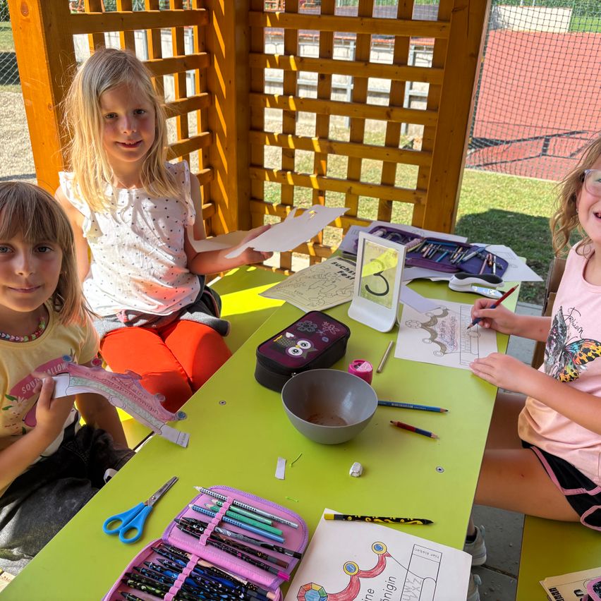 Drei Mädchen sitzen an einem grünen Tisch mit Malbüchern und Buntstiften. Der Tisch ist mit verschiedenen Bastelutensilien gefüllt. Die Mädchen scheinen an einer Malaktivität beteiligt zu sein.