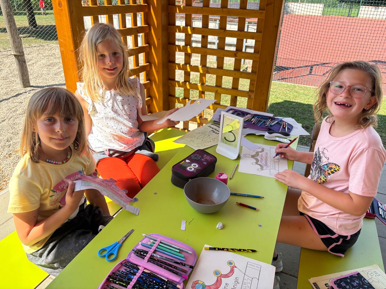 Drei Mädchen sitzen an einem grünen Tisch mit Malbüchern und Buntstiften. Der Tisch ist mit verschiedenen Bastelutensilien gefüllt. Die Mädchen scheinen an einer Malaktivität beteiligt zu sein.