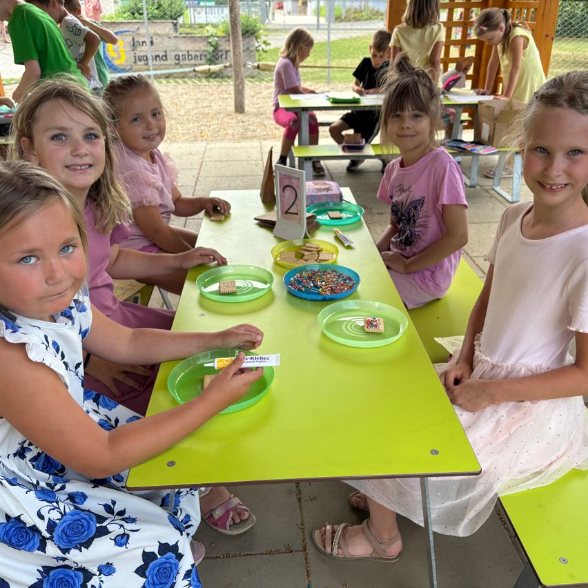 Fünf junge Mädchen sitzen an einem Tisch, lächeln und posieren für ein Foto. Der Tisch hat Teller mit Essen und ein Schild mit der Nummer 2. Dahinter sitzen andere Kinder, und ein Garten ist sichtbar.