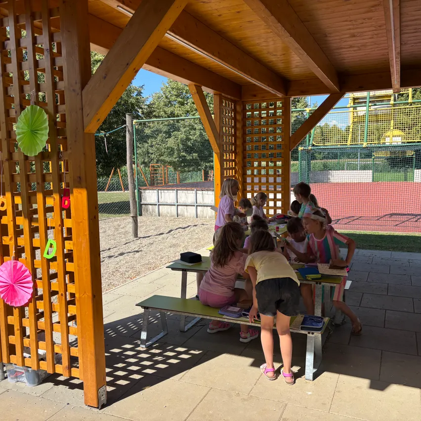 Eine Gruppe junger Mädchen sitzt an einem Picknicktisch unter einem hölzernen Pavillon, einige stehen und schauen nach unten. Sie scheinen in einer Aktivität verstrickt zu sein, möglicherweise einem Picknick. Der Pavillon ist mit Holzgittern und bunten Papierdekorationen verziert. Im Hintergrund befindet sich ein Spielplatz mit Netzzaun.