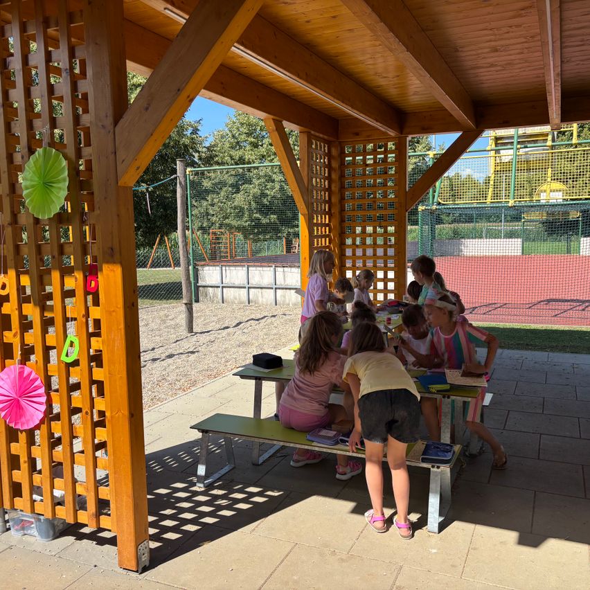 Eine Gruppe junger Mädchen sitzt an einem Picknicktisch unter einem hölzernen Pavillon, einige stehen und schauen nach unten. Sie scheinen in einer Aktivität verstrickt zu sein, möglicherweise einem Picknick. Der Pavillon ist mit Holzgittern und bunten Papierdekorationen verziert. Im Hintergrund befindet sich ein Spielplatz mit Netzzaun.