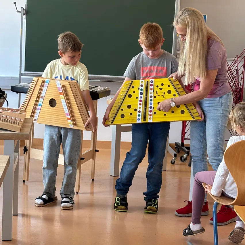 Zwei junge Jungen halten ein großes gelbes Instrument mit einer Frau hinter ihnen. Sie sind in einem Klassenzimmer mit einer Tafel und anderen Musikinstrumenten.