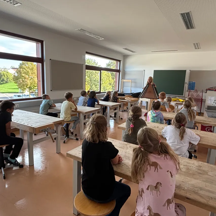 Ein Klassenzimmer mit Schülern an hölzernen Tischen, eine Lehrerin steht vor einer Tafel. Der Raum hat große Fenster mit Blick auf Bäume und ein Grasfeld.