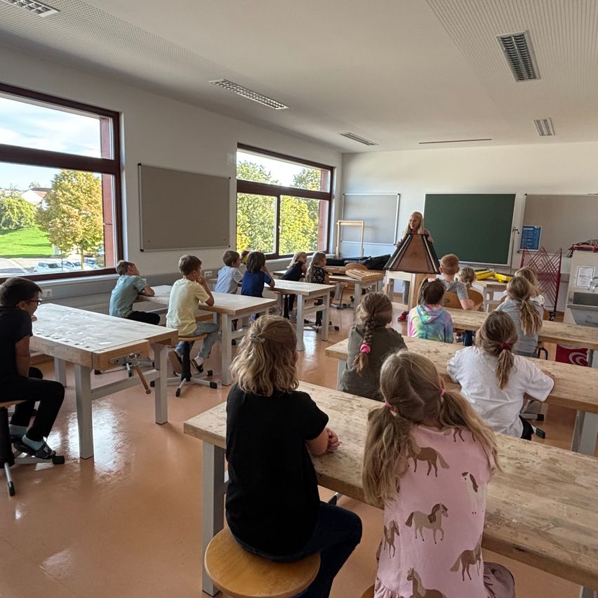 Ein Klassenzimmer mit Schülern an hölzernen Tischen, eine Lehrerin steht vor einer Tafel. Der Raum hat große Fenster mit Blick auf Bäume und ein Grasfeld.