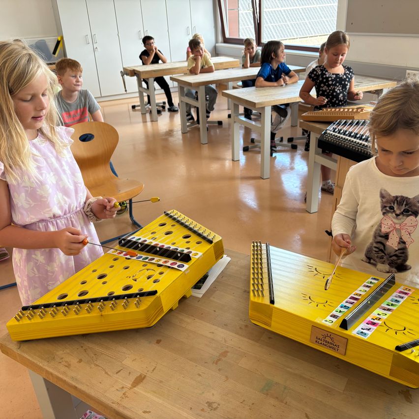 Zwei junge Mädchen spielen ein gelbes Xylophon in einem Klassenzimmer, während andere Kinder an Tischen sitzen.