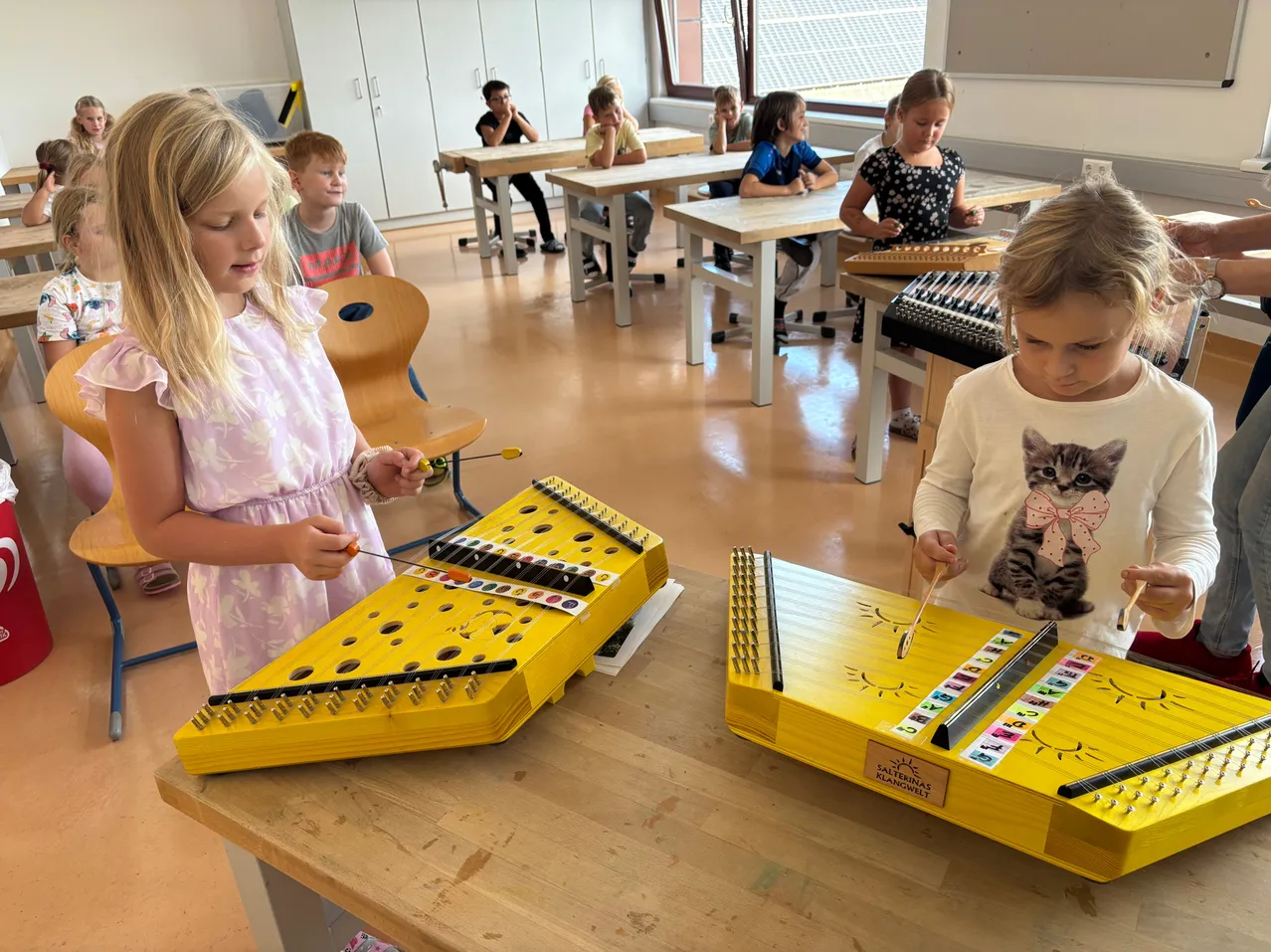 Zwei junge Mädchen spielen ein gelbes Xylophon in einem Klassenzimmer, während andere Kinder an Tischen sitzen.