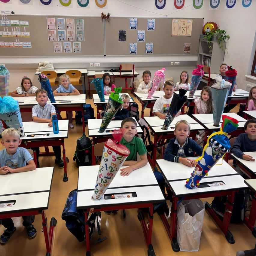 Ein Klassenzimmer voller junger Schüler, die an ihren Schreibtischen sitzen, jeder mit einem dekorierten Stiftetui vor sich. Die Klassenzimmerwände sind mit verschiedenen pädagogischen Postern und Dekorationen geschmückt.