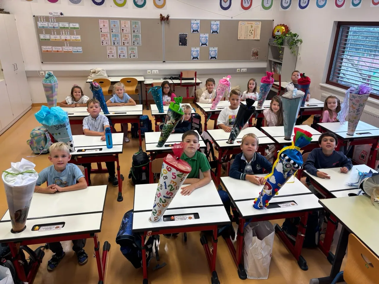 Ein Klassenzimmer voller junger Schüler, die an ihren Schreibtischen sitzen, jeder mit einem dekorierten Stiftetui vor sich. Die Klassenzimmerwände sind mit verschiedenen pädagogischen Postern und Dekorationen geschmückt.