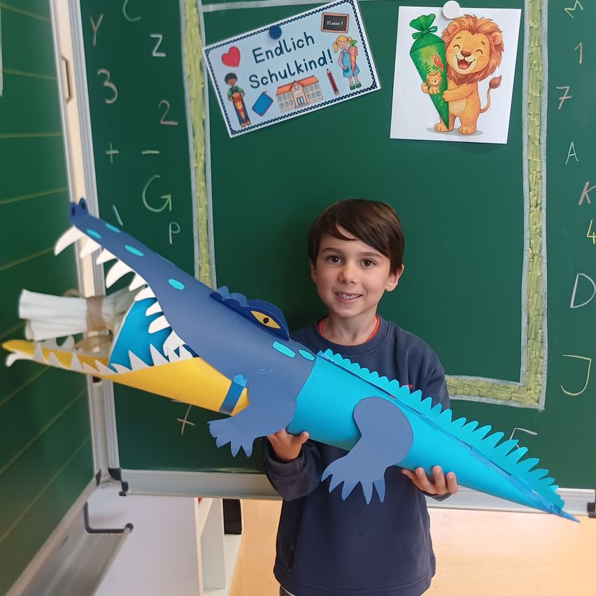 Ein junger Junge hält einen blauen Papierkrokodil vor einer Klassentafel. Die Tafel hat eine deutsche Botschaft, 'Endlich Schulkind!' und ein Bild eines Löwen.