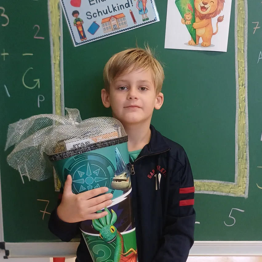Ein junger Junge hält einen großen, bunten Geschenkbeutel mit Drachendesign vor einer grünen Tafel.