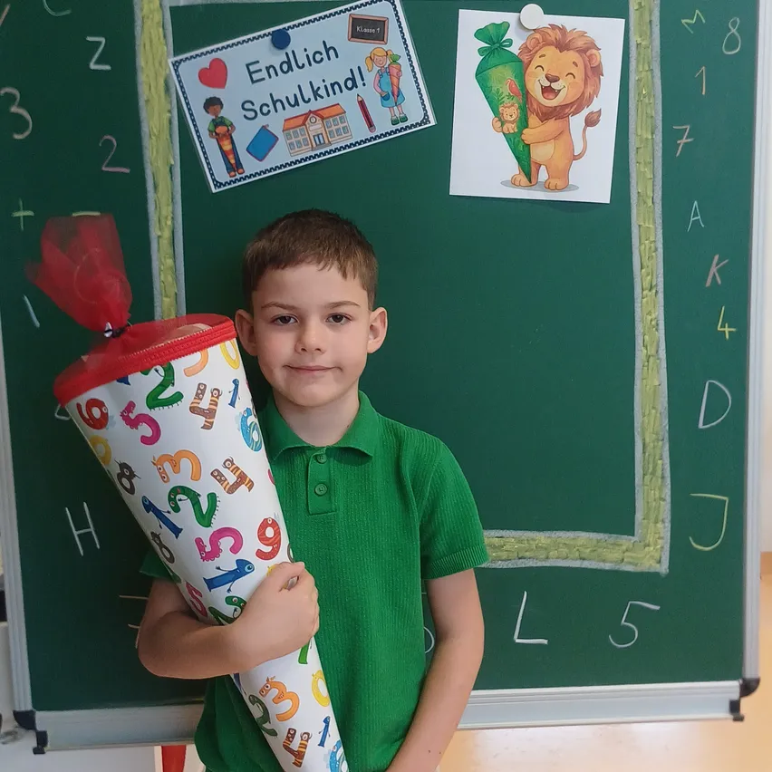 Ein junger Junge in einem grünen Shirt hält einen bunten Kegel mit Zahlen und Buchstaben und lächelt vor einer Klassentafel.
