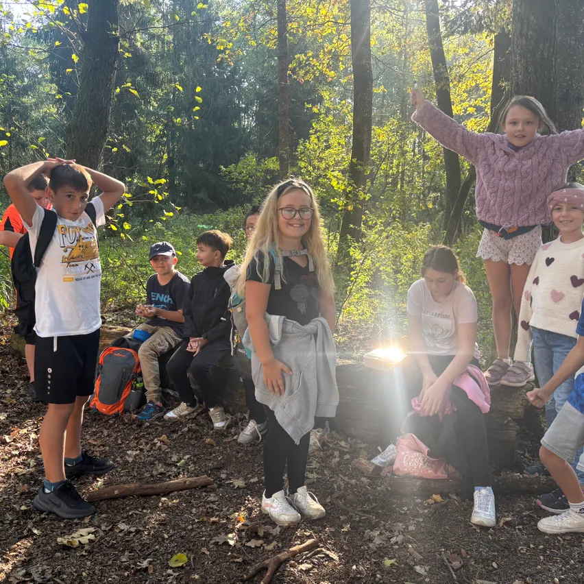 Eine Gruppe von Kindern steht und sitzt im Wald. Einige tragen Brillen und Turnschuhe.