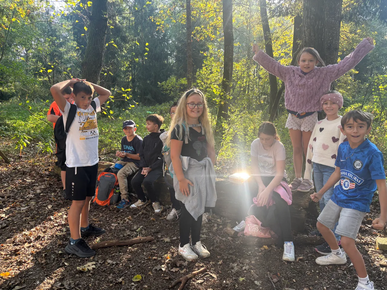 Eine Gruppe von Kindern steht und sitzt im Wald. Einige tragen Brillen und Turnschuhe.