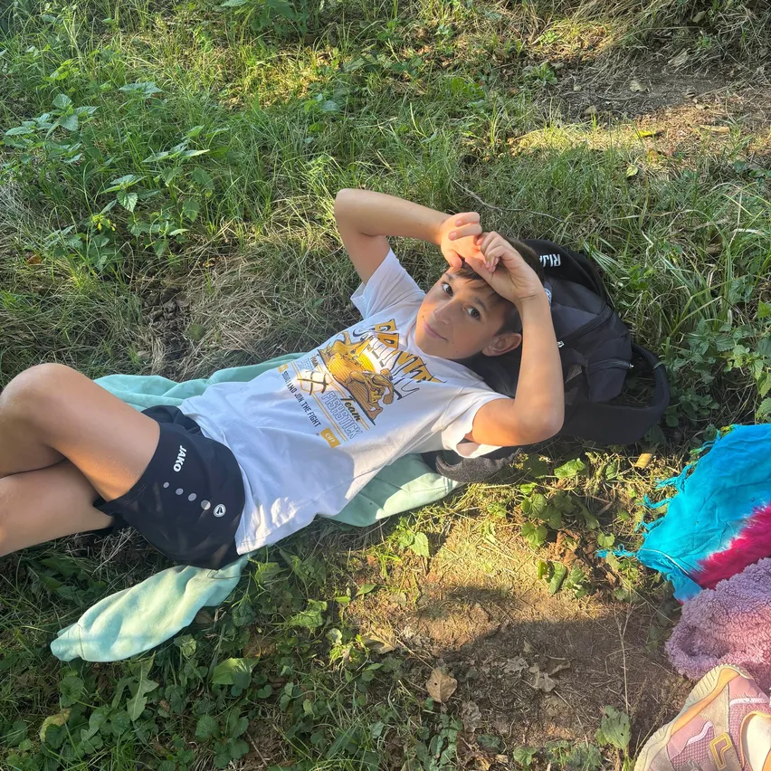 Ein junger Junge liegt auf einem Grasfeld, trägt ein weißes T-Shirt mit grafischem Design, schwarze Shorts und Turnschuhe. Ein Rucksack und ein Handtuch liegen in der Nähe.
